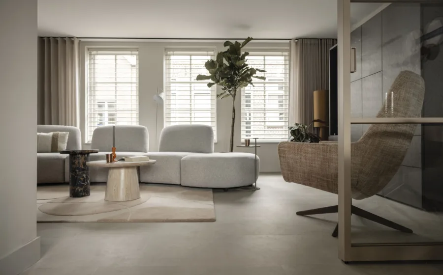 Woonkamer uit vtwonen Weer Verliefd op je Huis, Grafhorst, met lichtgrijze hoekbank, ronde travertin salontafel, gevlochten fauteuil en een grote vijgenboom voor de vensterbank.