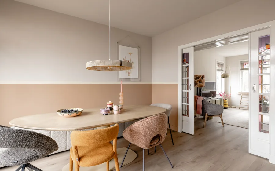 Eetkamer met verf lambrisering en ovale tafel met stoelen in verschillende kleuren.