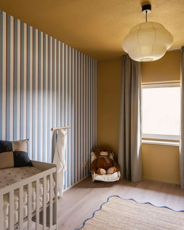 Schattige babykamer met streepjesbehang, okergele en blauwe accenten, Loft79 gordijnen in Wilg 20 en kleine accessoires die een vrolijke en knusse sfeer creëren.