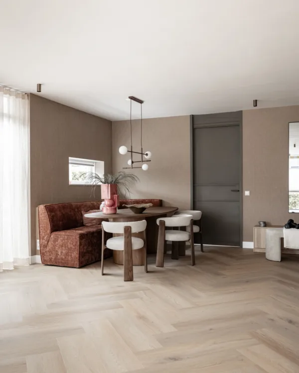 Eethoek met een lichte eiken visgraatvloer in een warme, natuurlijke kleur. Op de vloer staan een ronde houten tafel met crèmekleurige stoelen en een roestkleurige hoekbank, tegen taupe muren.