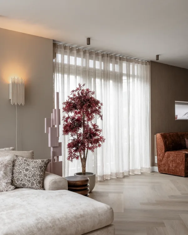 Ruime woonkamer met transparante beige linnen gordijnen met golfplooi voor een hoog raam. Ze laten zacht daglicht door en passen bij de warme aardetinten van de muren, de bank en de eethoek.