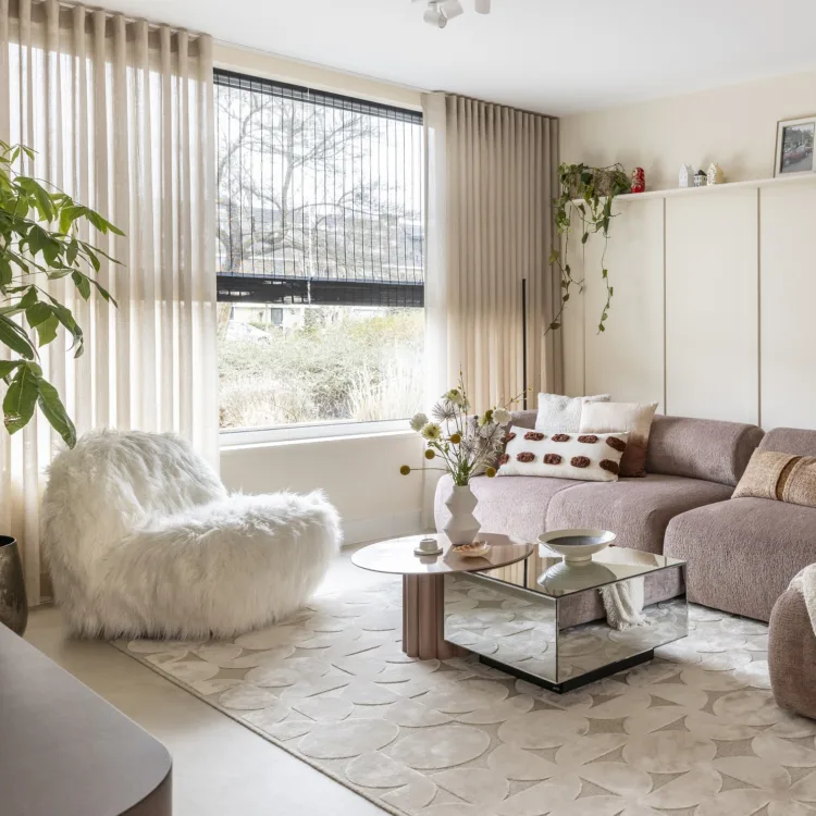 Woonkamer met betonlook vinylvloer en zacht vloerkleed, sfeervolle inbetween gordijnen en zwarte geweven hout vouwgordijnen.