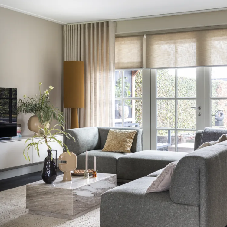 Woonkamer met beige rolgordijnen en overgordijnen, grijze hoekbank en marmer look tafeltje