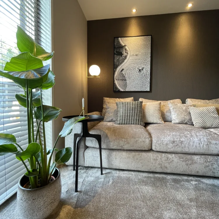 Hotel chique woonkamer met beige loungebank en sierkussens, donkere accentmuur met kunst, zwarte bijzettafel, warme wandlamp en grote groene kamerplant bij het raam.