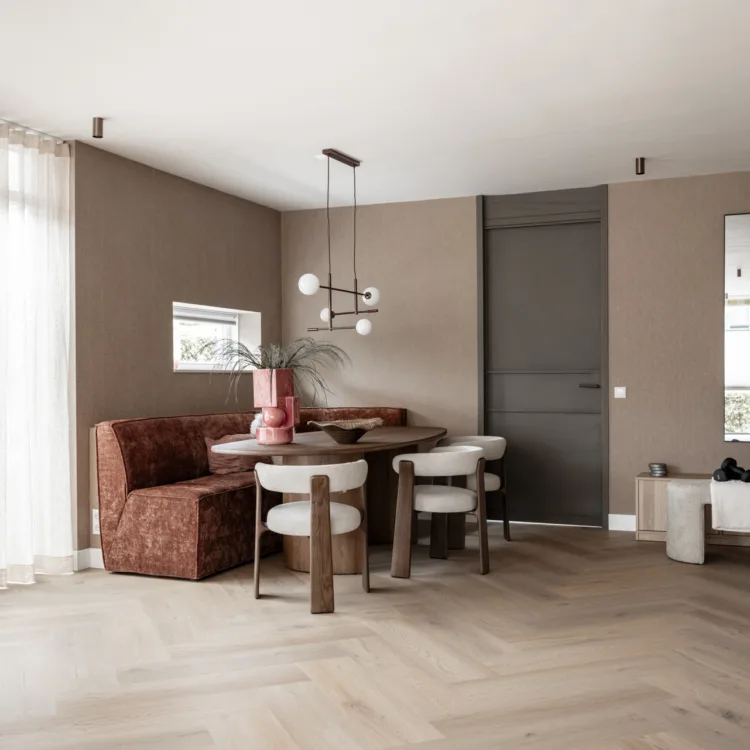 Eethoek met een lichte eiken visgraatvloer in een warme, natuurlijke kleur. Op de vloer staan een ronde houten tafel met crèmekleurige stoelen en een roestkleurige hoekbank, tegen taupe muren.