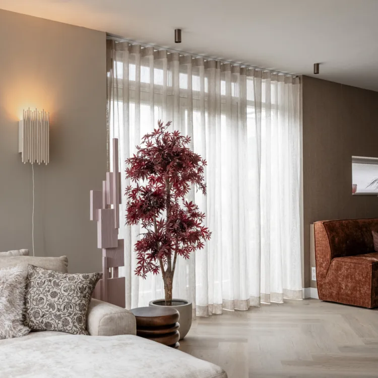 Ruime woonkamer met transparante beige linnen gordijnen met golfplooi voor een hoog raam. Ze laten zacht daglicht door en passen bij de warme aardetinten van de muren, de bank en de eethoek.
